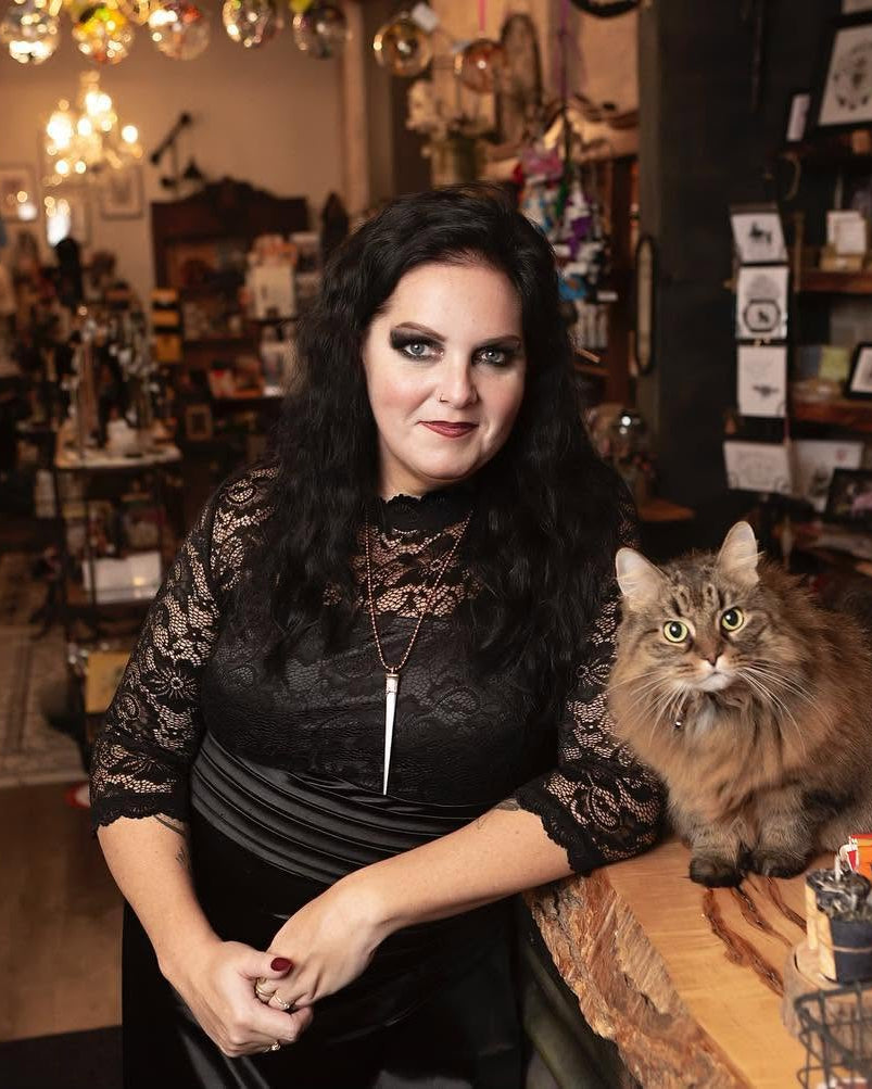 A woman with long black hair wearing a black lace top stands behind a wooden counter in a cluttered vintage shop, with a fluffy brown tabby cat sitting on the counter next to her.