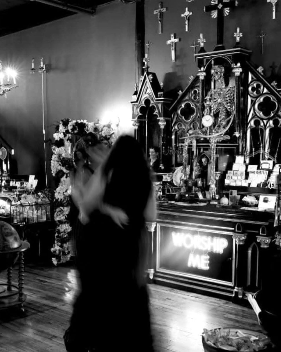 A black and white photograph of a gothic shop interior, featuring a blurred figure in the foreground, ornate altars, crosses on the walls, and display cases filled with items.
