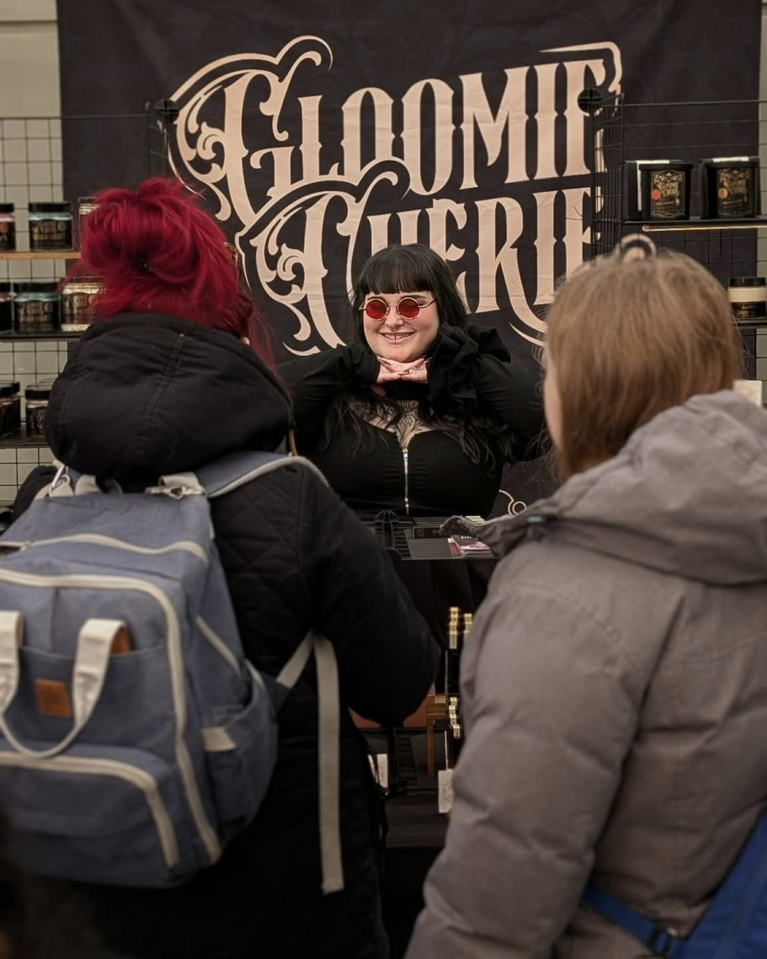 Person in a black outfit with sunglasses sitting at a table in front of a 'Gloomie Ghorie' banner, with two people facing them.