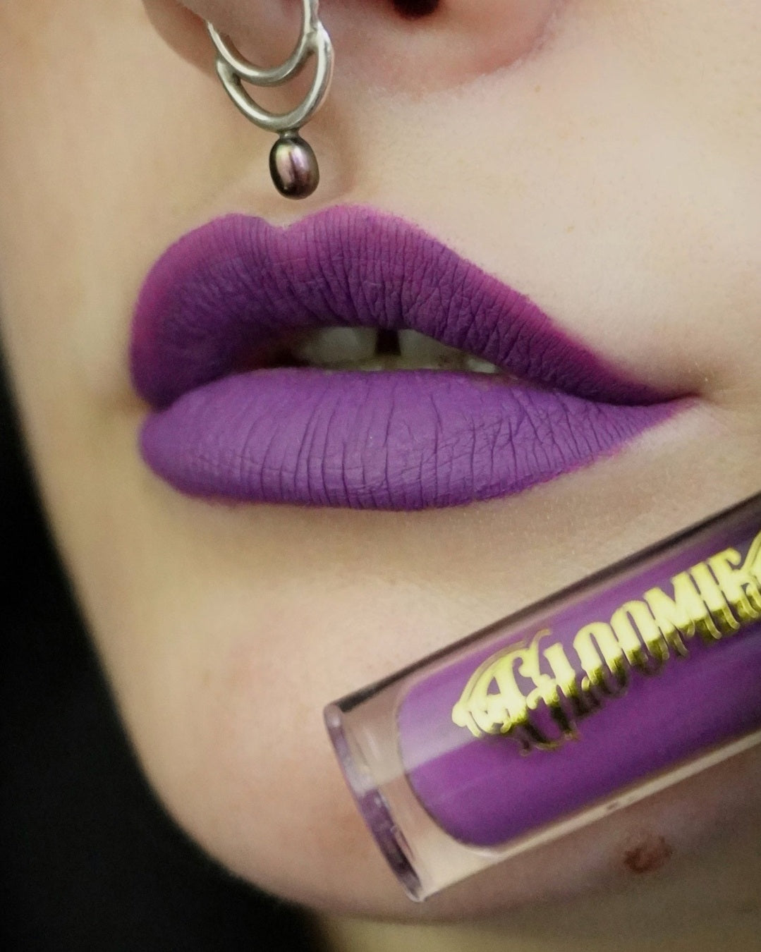 Close-up of a person wearing purple lipstick with a lip gloss tube in the foreground.