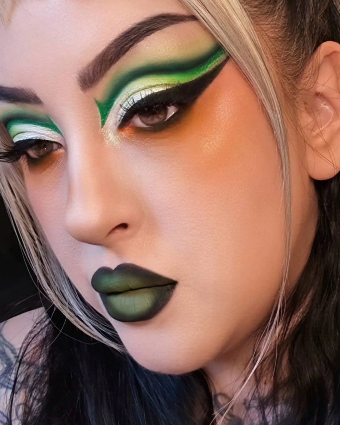 Close-up of a person with dramatic green and black eye makeup, olive green lipstick, and styled hair against a dark background.