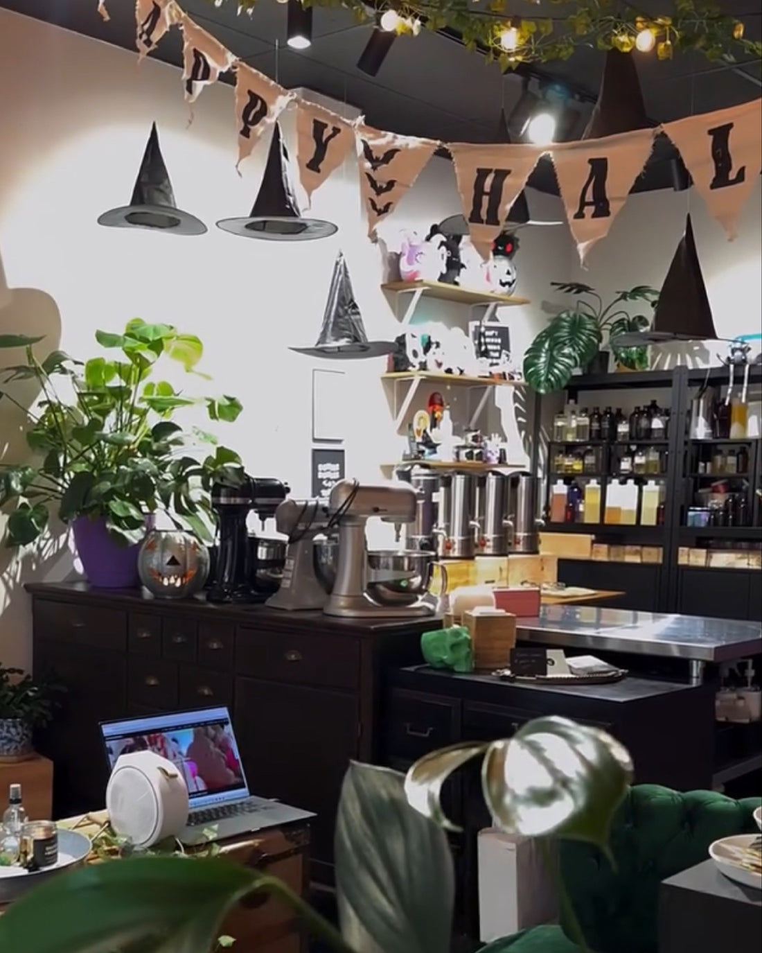 A cozy, decorated cafe interior with Halloween-themed bunting, witch hats, potted plants, and a dark wood counter. A laptop is open on a table in the foreground, and text at the bottom reads 'It was always Halloween at the shop'.
