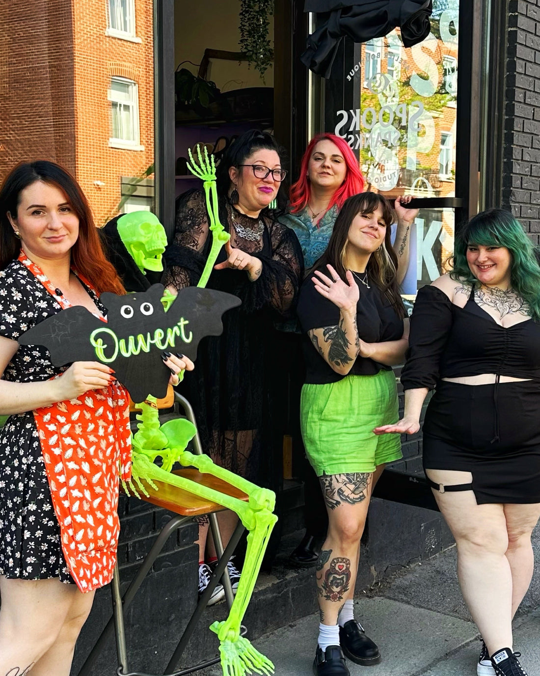 A group of six women posing in front of a storefront on a city sidewalk, dressed in Halloween costumes. One woman holds a black bat-shaped sign that reads 'Ouvert' next to a large green skeleton decoration.